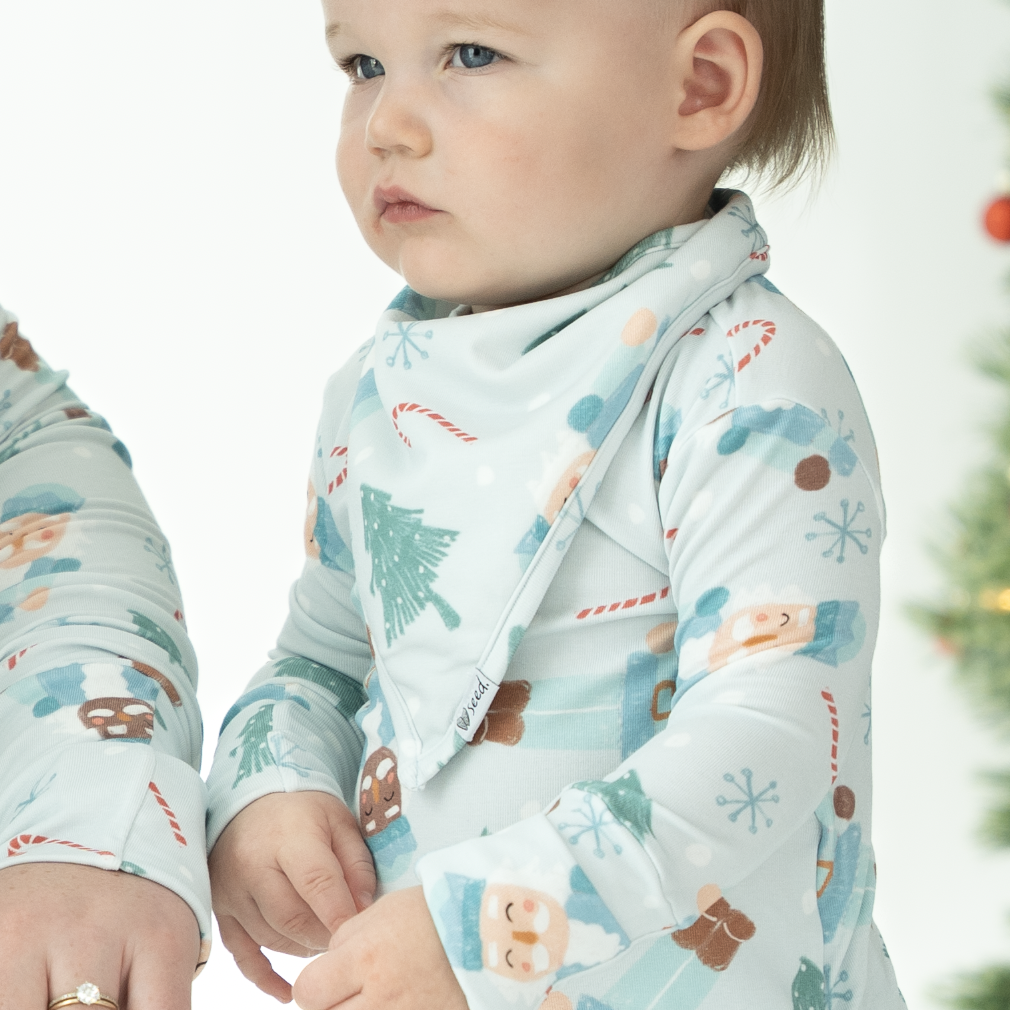 A baby wearing a long-sleeve light blue holiday-themed sleep sack with a bib-style neckline, standing next to an adult arm. The background features a softly blurred Christmas tree with warm lights.
