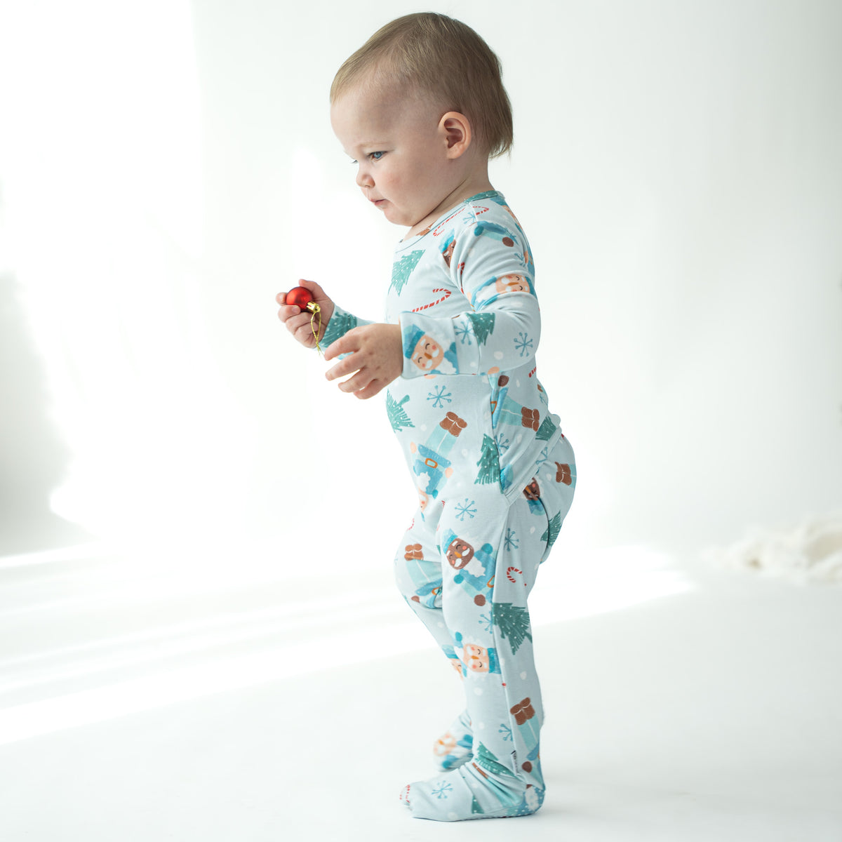 A baby standing in profile wearing a light blue footed one-piece pajama with a Christmas print of houses, trees, and snowflakes, holding a small red toy in a bright white room.