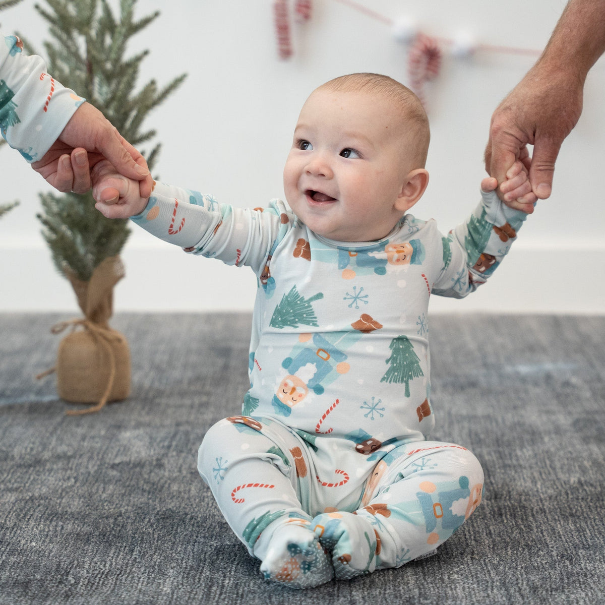 A baby sitting on a gray carpet holding hands with two adult hands, wearing a light blue footed sleeper with a Christmas tree and snowman pattern. A small potted pine tree and a candy cane garland are in the background.
