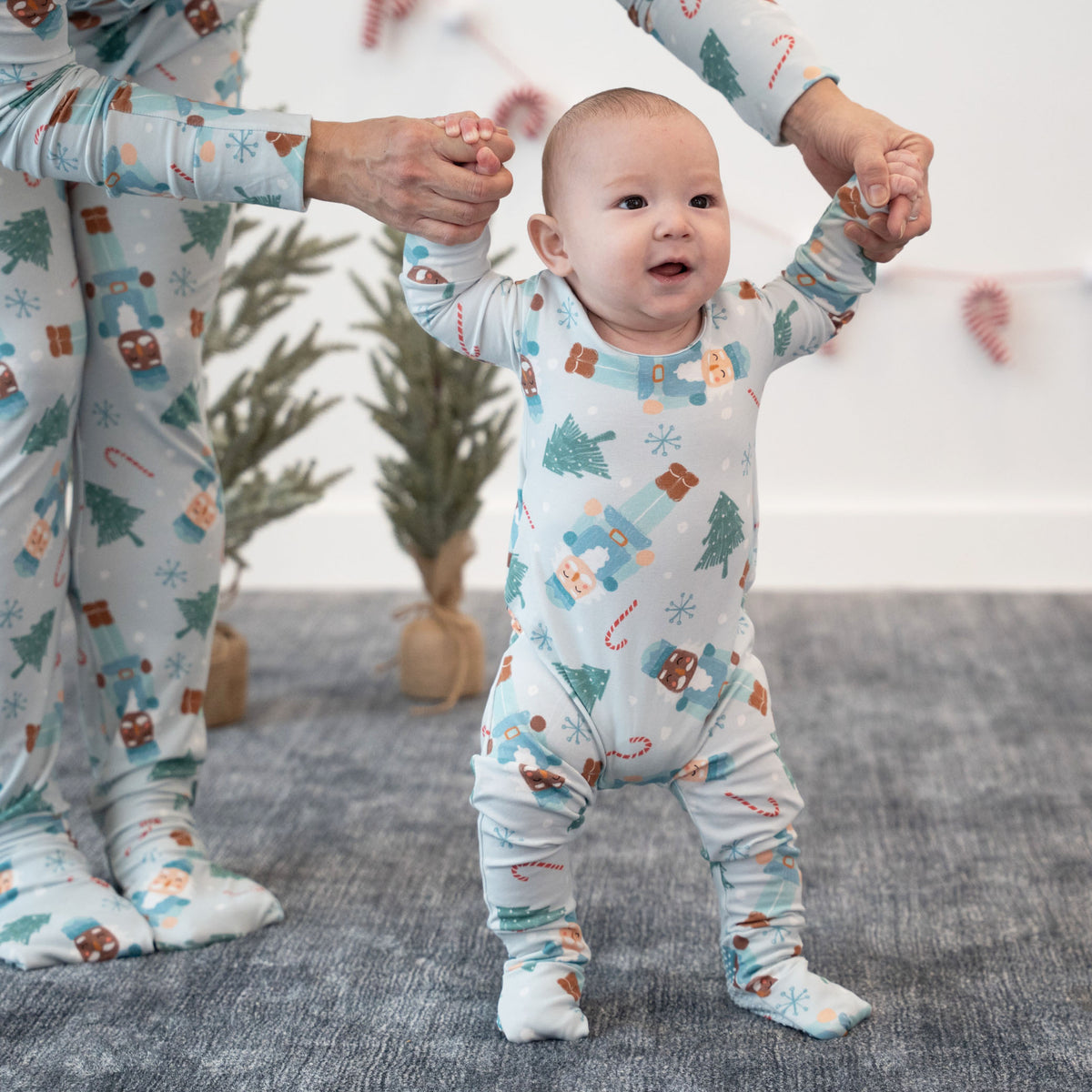 A baby wearing a light blue footed sleeper with a holiday gingerbread print is learning to walk while being held up by an adult wearing matching pajamas. The scene is set on a gray textured rug with small potted trees in the background.