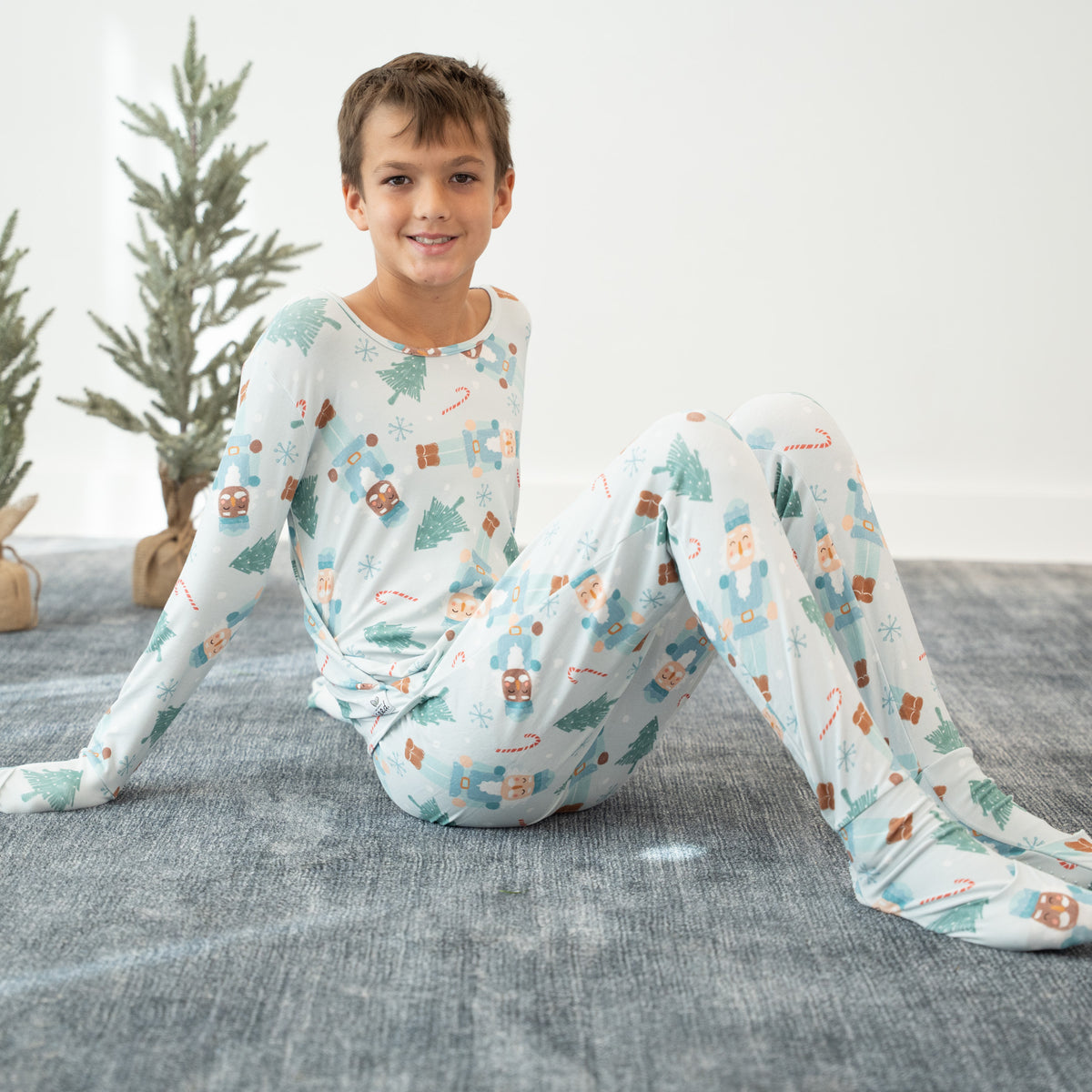 A young boy wearing a long-sleeve footed pajama set with a Christmas-themed print of gingerbread men, candy canes, and pine trees, sitting on a gray rug.