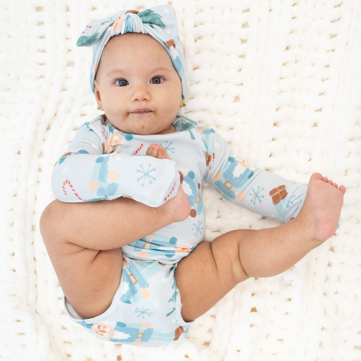 A baby lying on a white knit blanket, wearing a light blue long-sleeve bodysuit with a winter-themed print and a matching headband.
