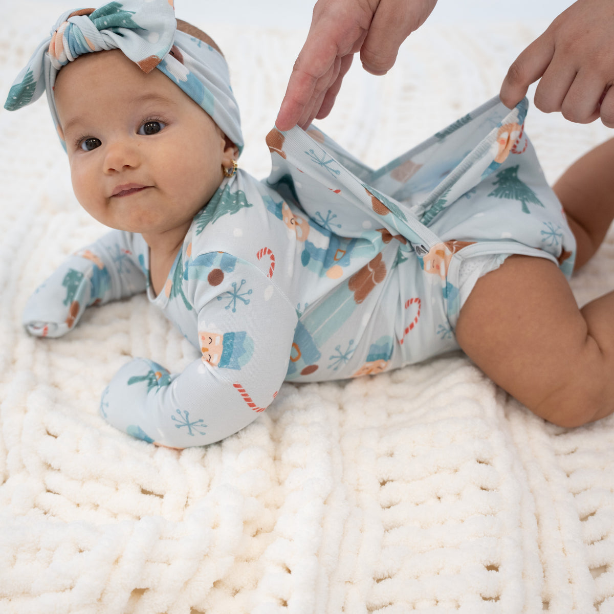 A baby lying on a white knit blanket wearing a light blue holiday print bodysuit and matching headband, while an adult's hands adjust the diaper area of the outfit.