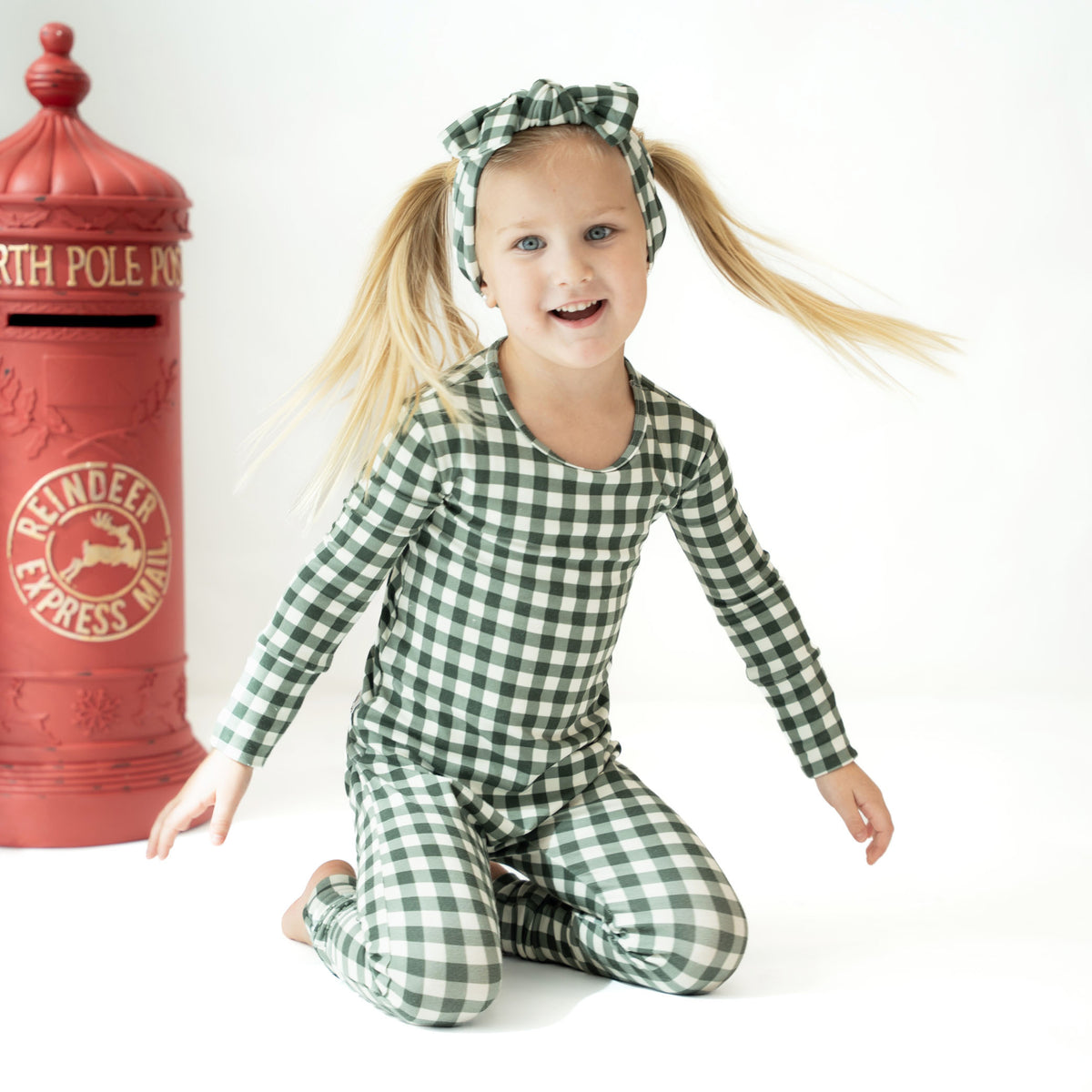 A young girl with blonde pigtails sits smiling on a white studio floor, wearing a green and white gingham long-sleeve pajama set with a matching headband. A red vintage-style North Pole Express Mail mailbox is visible to her left.
