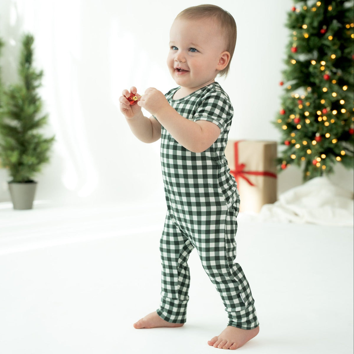 A baby wearing a green and white gingham check one-piece jumpsuit stands barefoot in a bright, festive room with a Christmas tree and wrapped gift in the background.