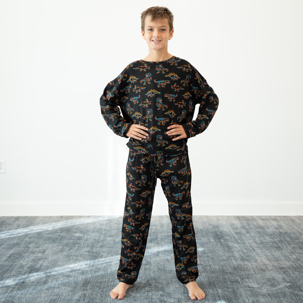 A child standing barefoot indoors wearing a black long-sleeve pajama top and matching pants with an allover colorful dinosaur print, posed with hands on hips against a white wall and gray floor.