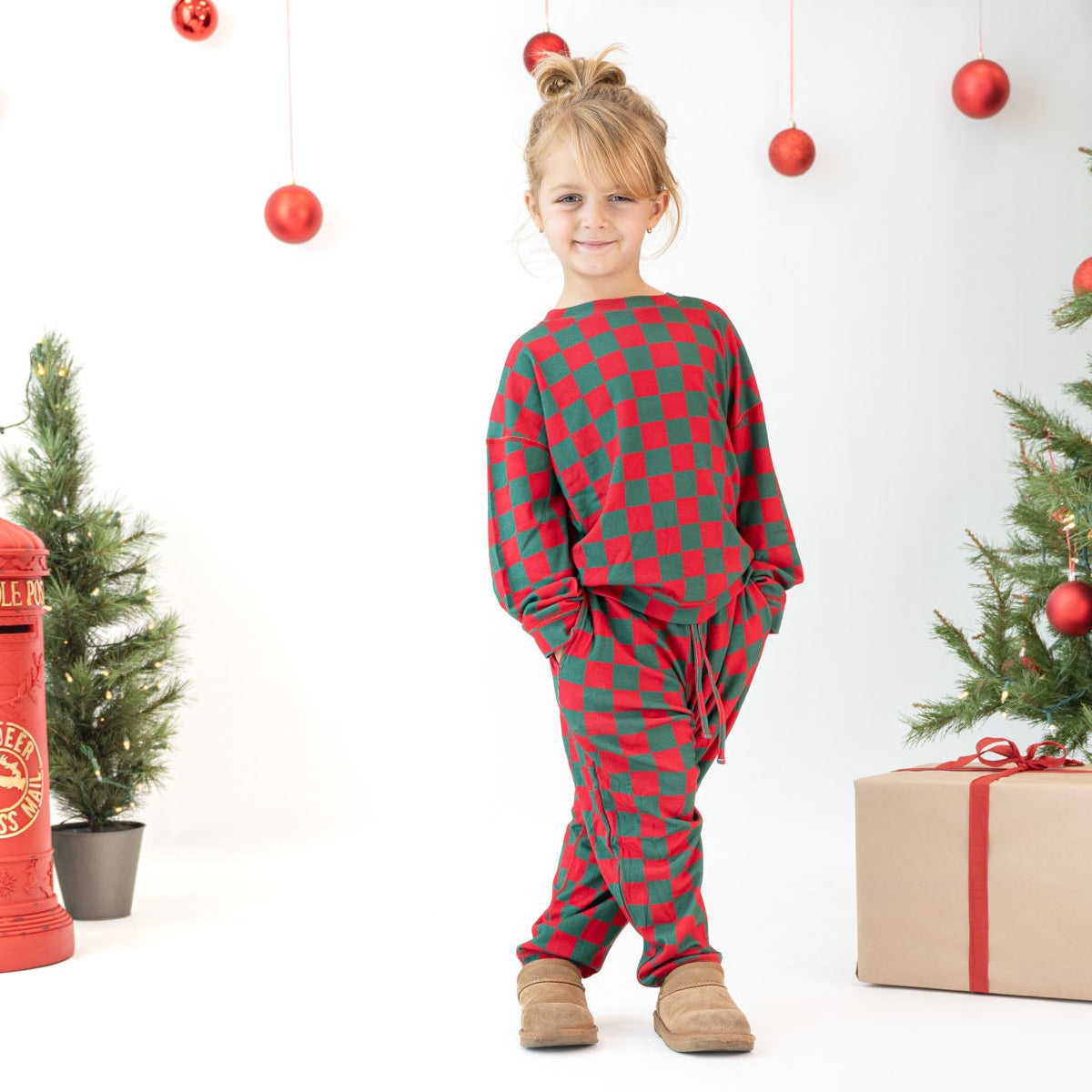 A young girl stands in a studio setting wearing a red and green plaid holiday pajama set. The background is decorated for Christmas with a tree, ornaments, wrapped gifts, and a red vintage post office prop.