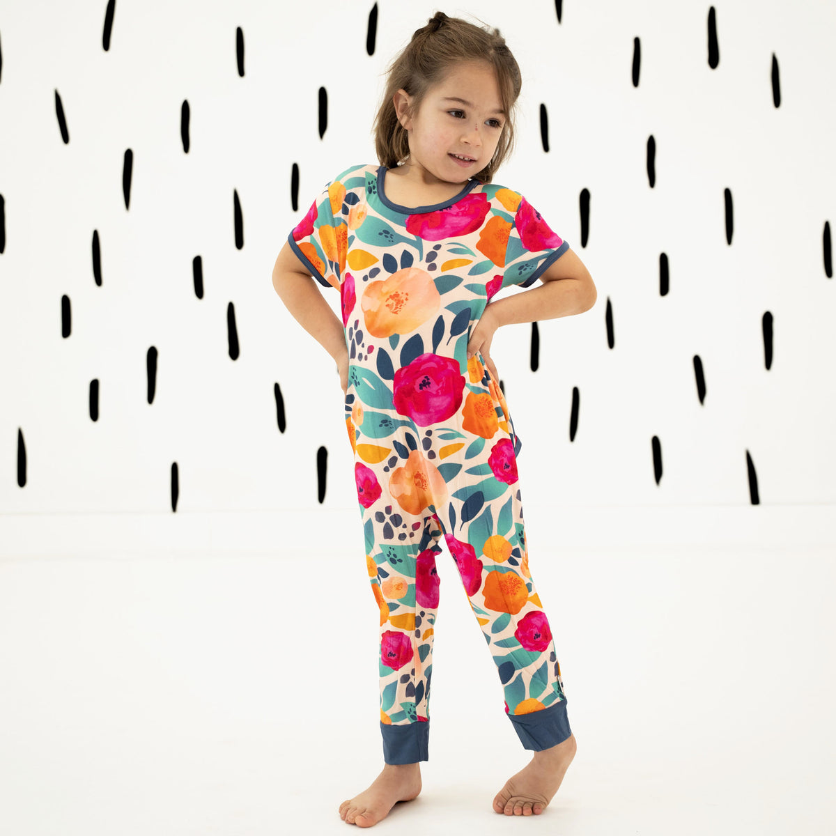 A young girl stands barefoot wearing a short-sleeve one-piece pajama with a colorful floral print. The pajama features a vibrant pattern of pink, orange, yellow, and teal flowers and leaves on a light background, with solid navy blue trim at the collar, sleeve cuffs, and ankle cuffs.