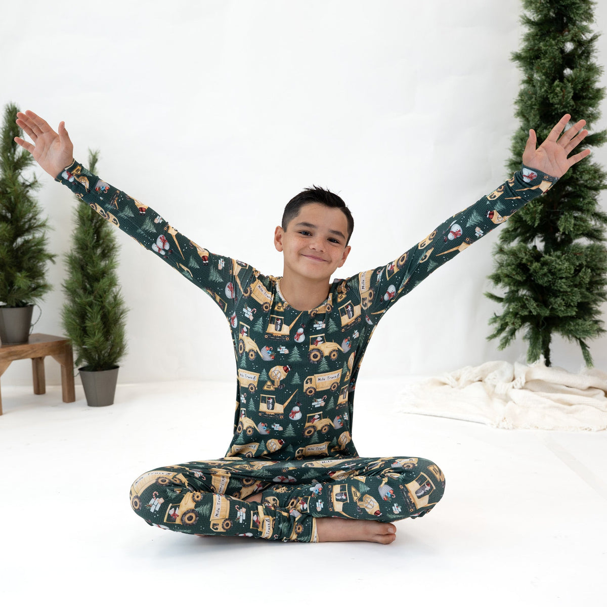 A young boy wearing a dark green long-sleeve pajama set with a cartoon construction truck pattern, sitting cross-legged with his arms raised in a studio setting decorated with small potted trees.
