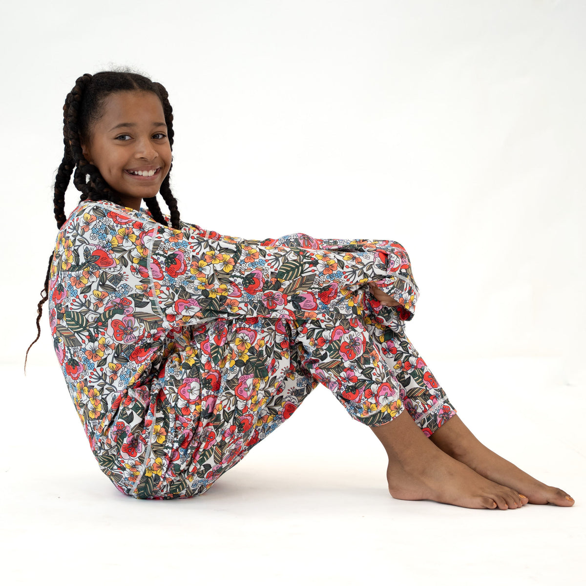 A young girl with braided hair sits cross-legged on a white studio background, wearing a loose floral jumpsuit with a multicolor print of red, pink, yellow, blue, and green flowers on a dark base.