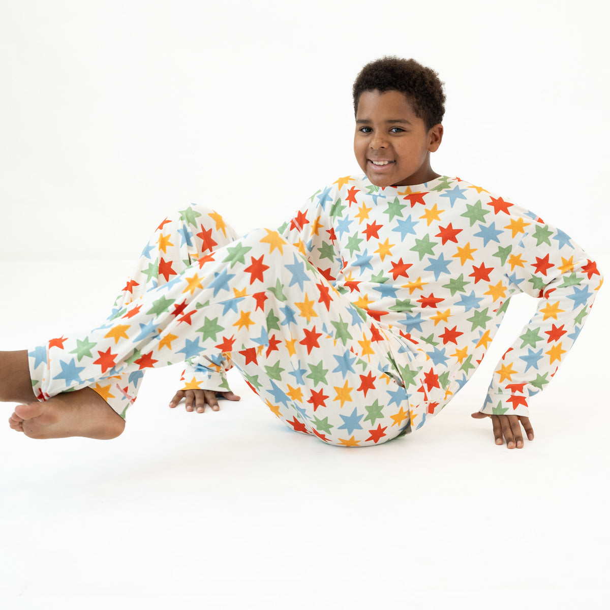 A young boy wearing a long-sleeve pajama set with a colorful star pattern, sitting on a white studio floor.