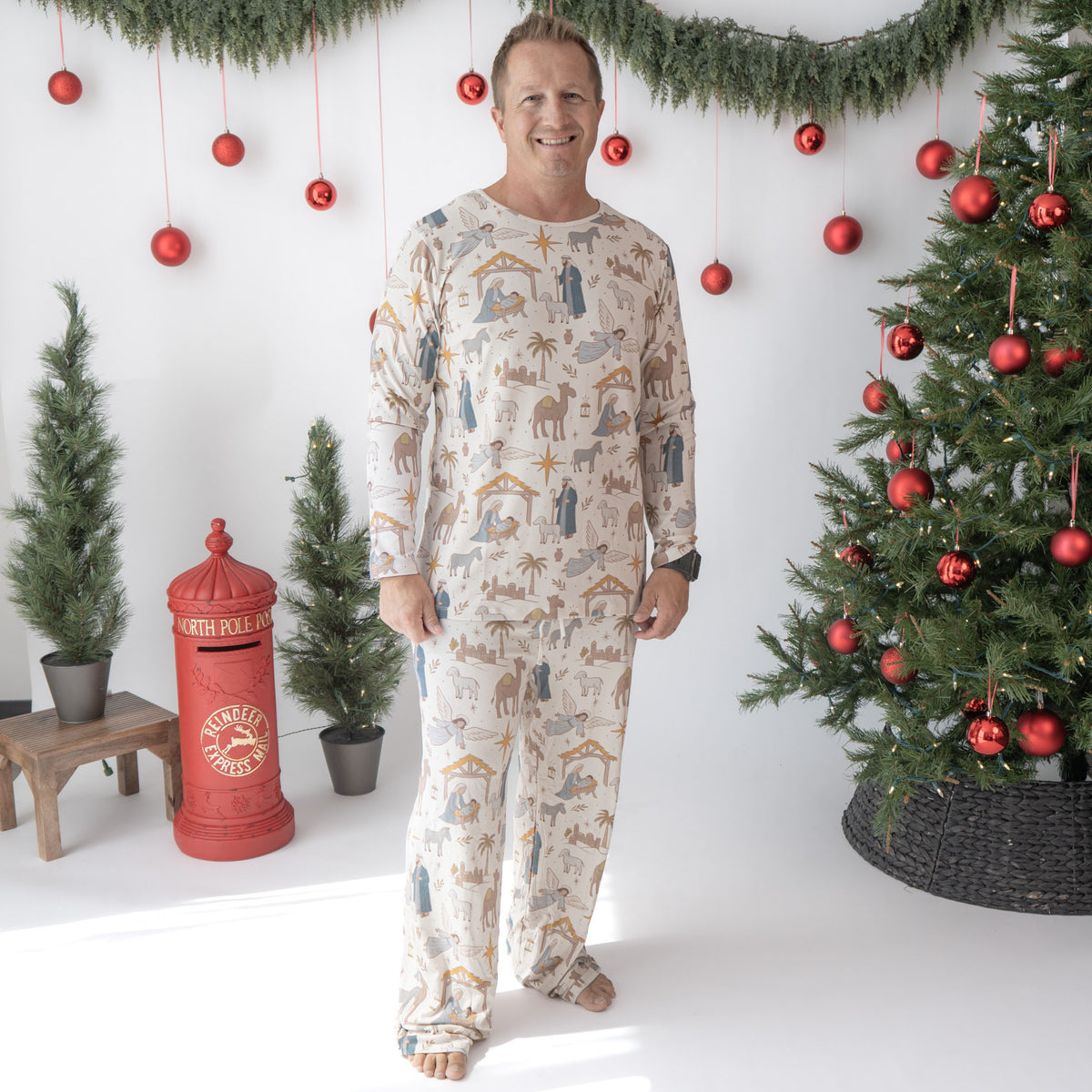 A man wearing a long-sleeve Christmas-themed pajama set with an all-over nativity scene print, standing in a decorated room with a Christmas tree, garland, and red ornaments.