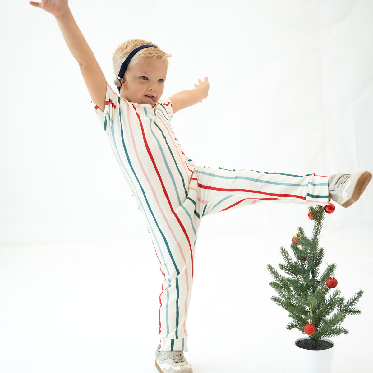 A young child wearing a festive holiday-themed striped jumpsuit with red, teal, and white vertical stripes, balancing on one foot next to a small decorated Christmas tree against a white studio background.