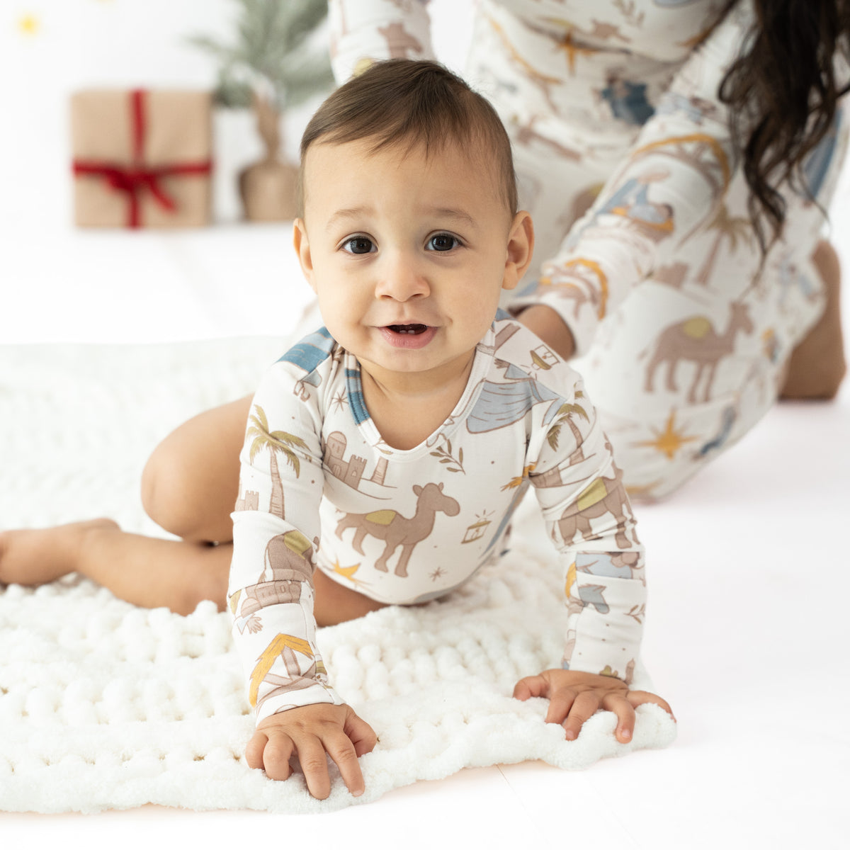 A baby crawling on a white textured blanket, wearing a long-sleeve cream pajama top with a safari animal print. An adult in matching print is visible in the background, along with a wrapped gift box and small plants.