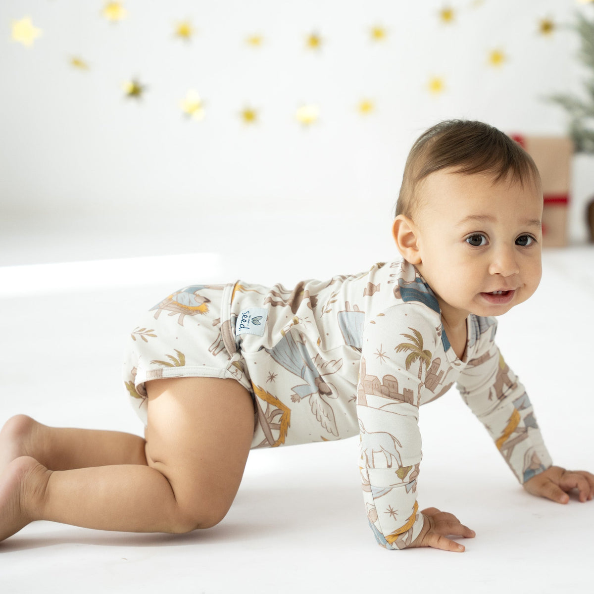 A baby crawling on a white floor wearing a long-sleeve cream onesie with a tropical print of palm trees, birds, and fish, set against a decorated white background with gold star garlands.