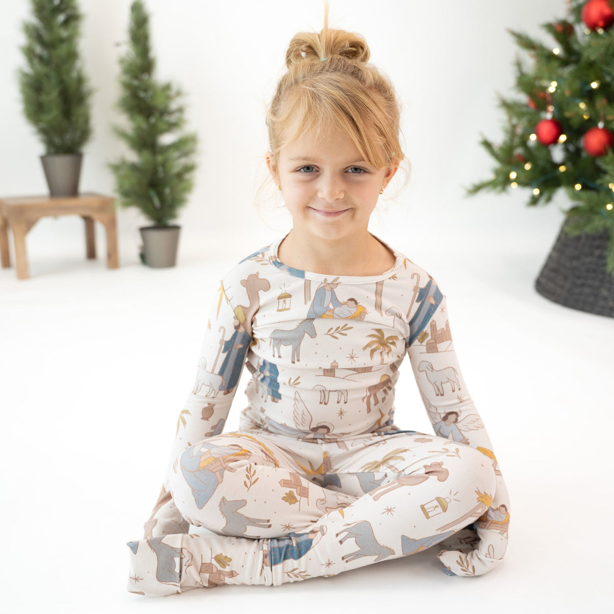 A young girl with blonde hair in a bun sits cross-legged on a white floor, wearing a matching long-sleeve pajama set with an animal print. The pajamas feature a cream base with illustrations of safari animals like elephants, giraffes, and zebras in muted blue, brown, and gold tones.