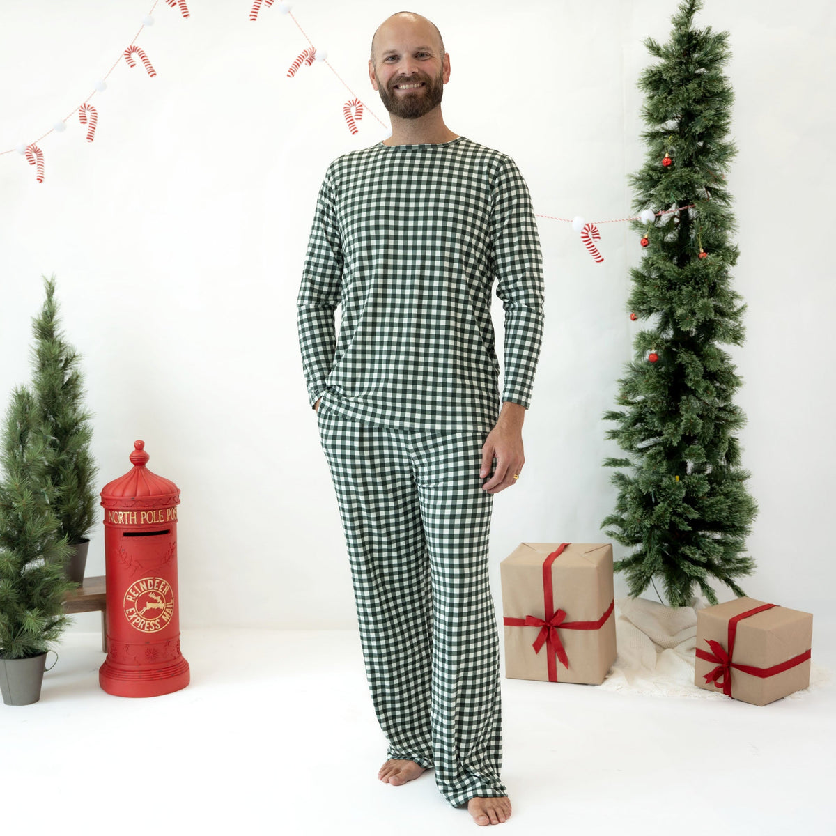 A man wearing a green and white gingham long-sleeve pajama set stands barefoot in a holiday-themed studio setting with Christmas trees, wrapped gifts, and a red post box.