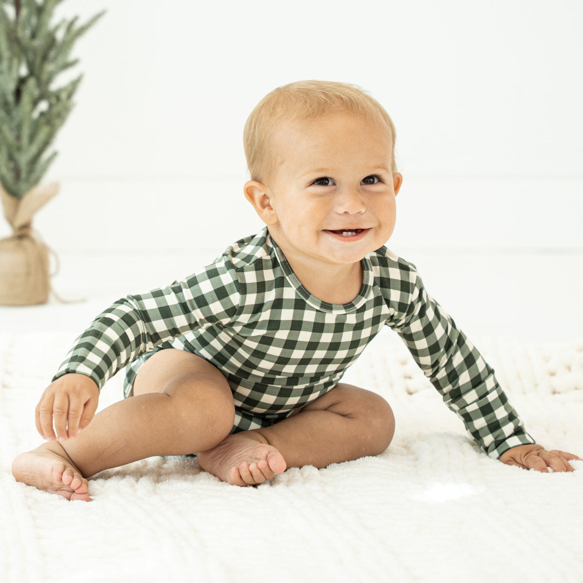 A smiling baby sitting on a white textured blanket, wearing a green and white gingham long-sleeve romper. A small potted tree is visible in the background against a white wall.