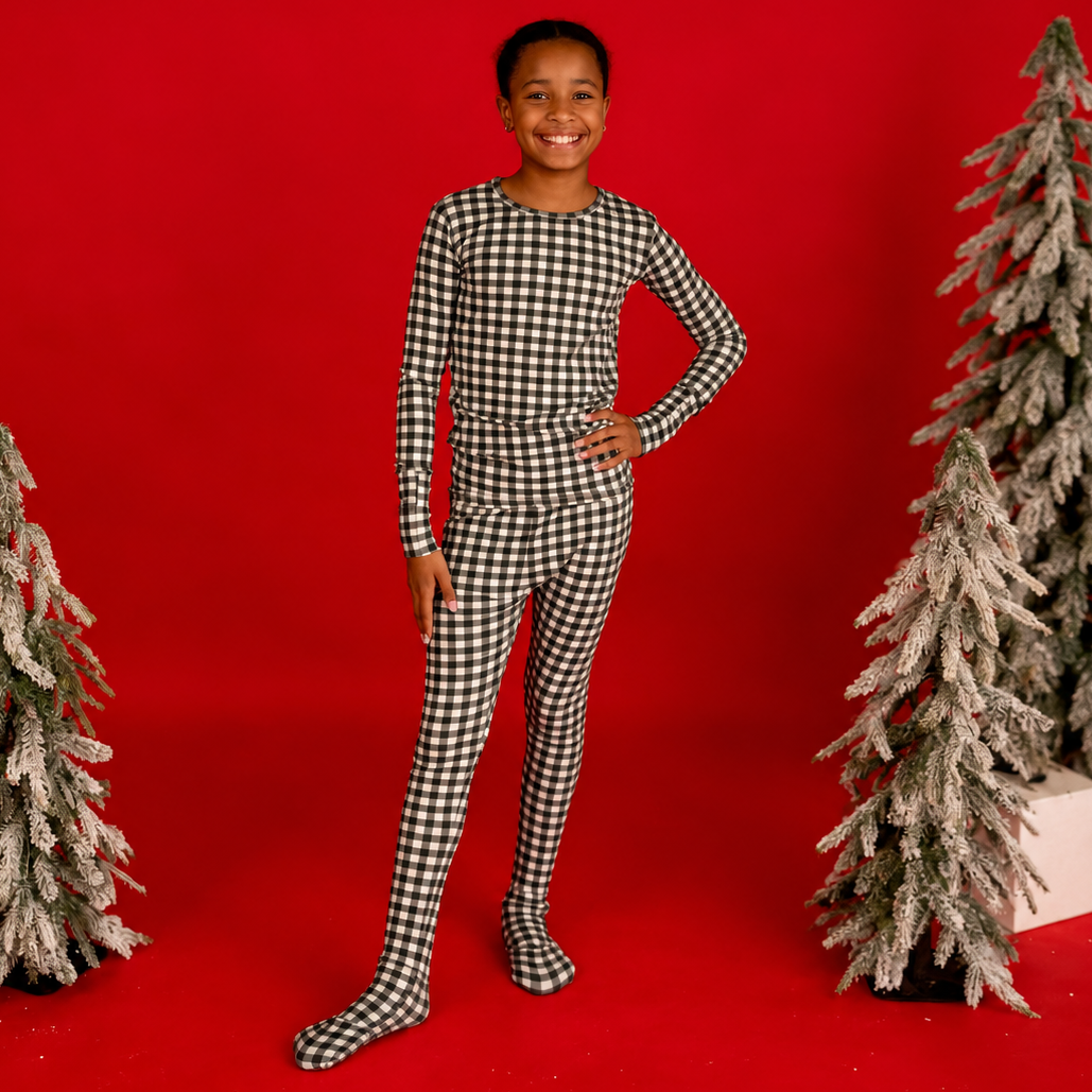 A child modeling a black and white gingham check long-sleeve tummy shirt and matching full-length leggings against a solid red studio backdrop, flanked by decorative flocked Christmas trees.