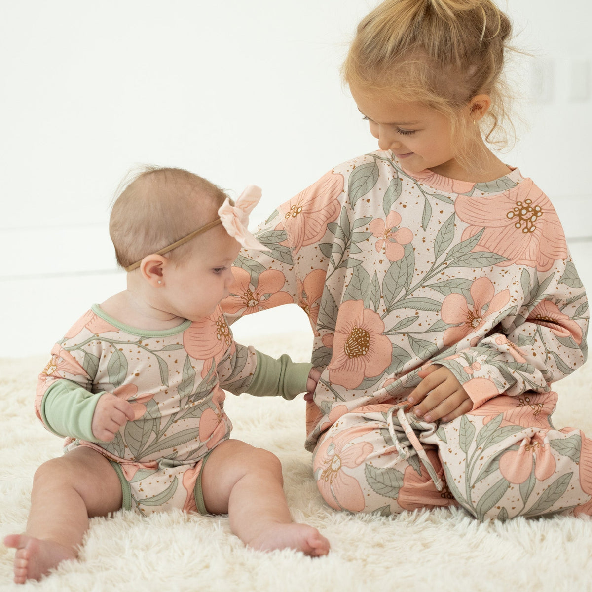 A toddler and baby sitting on a white fluffy rug, both wearing matching floral pajamas in soft pink, sage green, and beige tones.