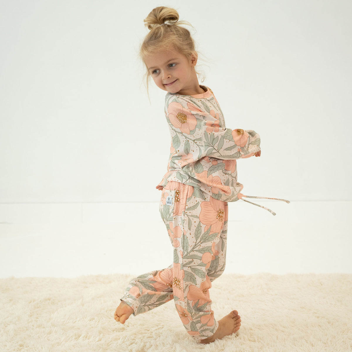 A young girl with blonde hair in a bun is wearing a two-piece pajama set with a floral print, standing on a white shag rug against a white studio background.