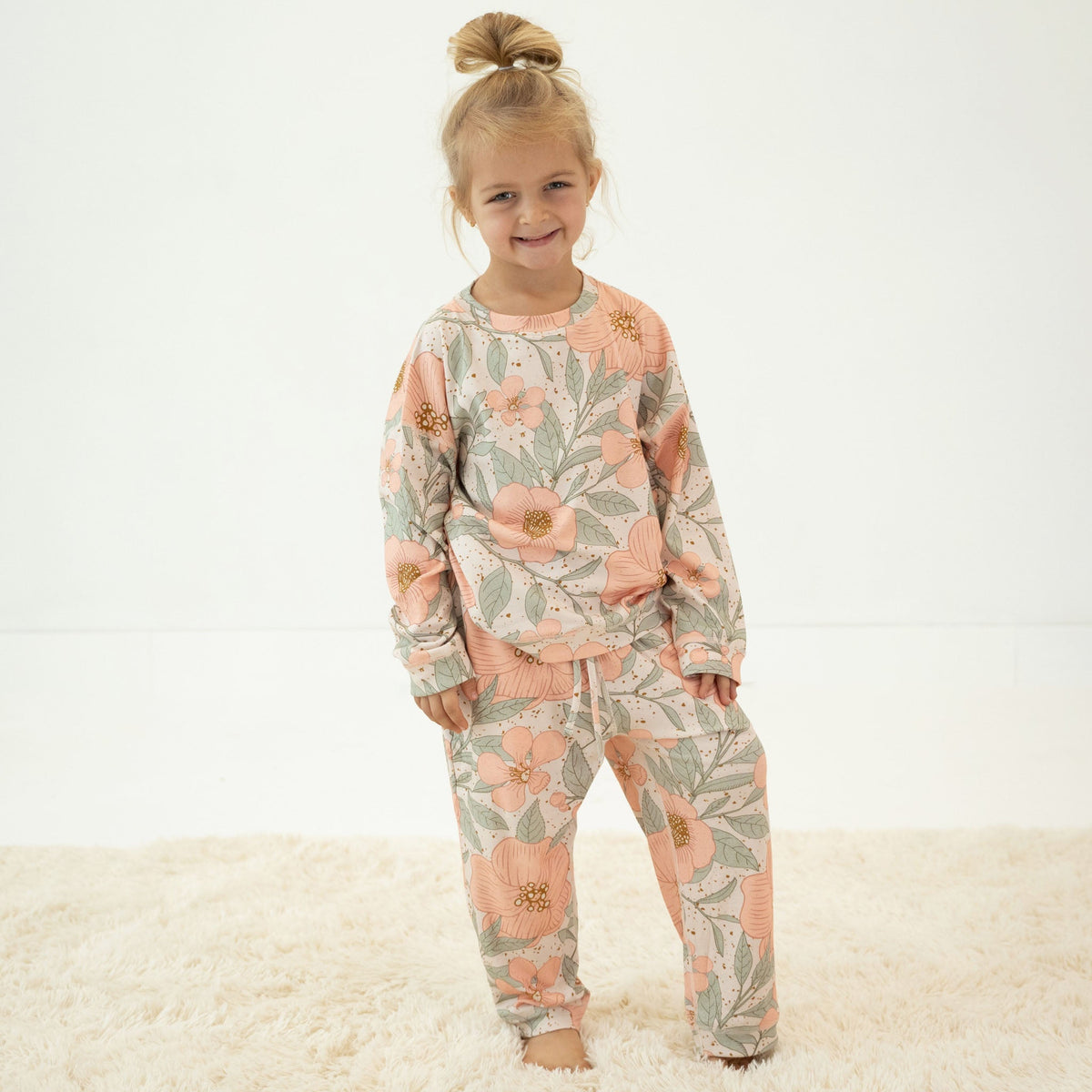 A young girl with blonde hair in a bun stands barefoot on a white shag rug, wearing a matching two-piece pajama set with a peach and sage green floral pattern.