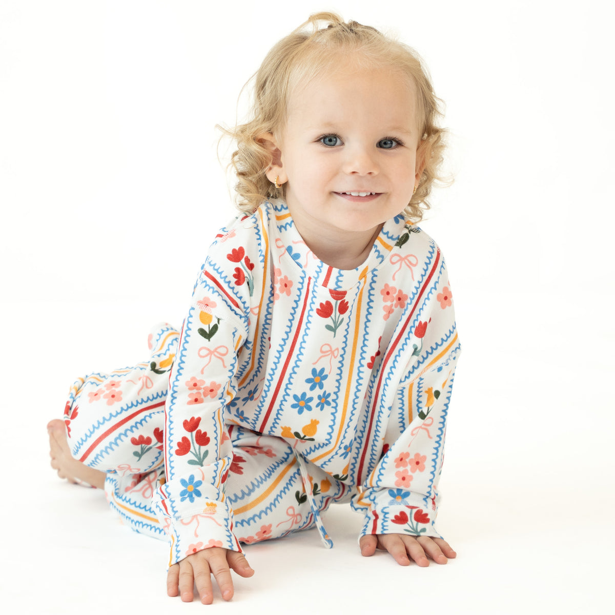 A smiling toddler with blonde curly hair sits on a white studio floor wearing a long-sleeve, one-piece baby/toddler outfit with a colorful floral and geometric pattern.