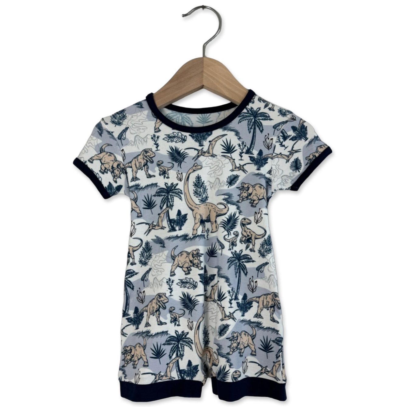 A short-sleeved, short-legged romper for kids, featuring a dinosaur print, hanging on a wooden hanger.