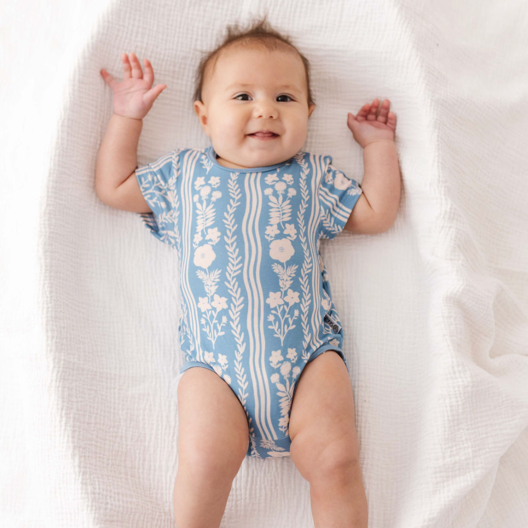 Baby lying on a white blanket wearing a blue and white patterned onesie.