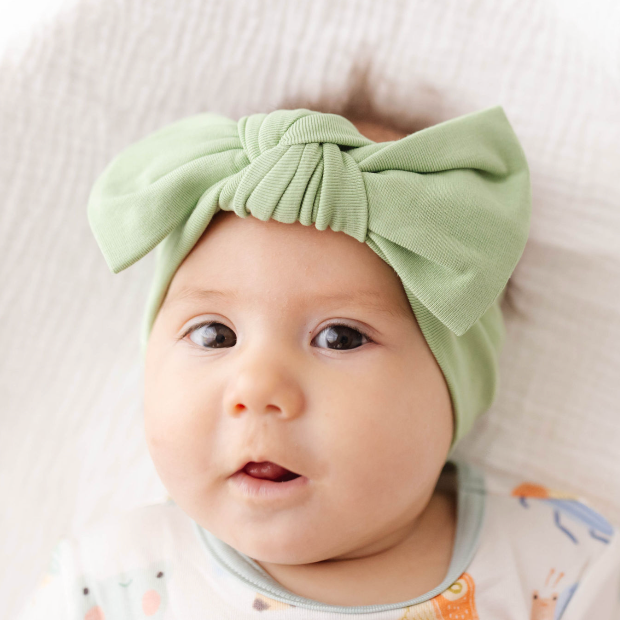 Baby wearing a green headband with a bow, sitting on a white surface.