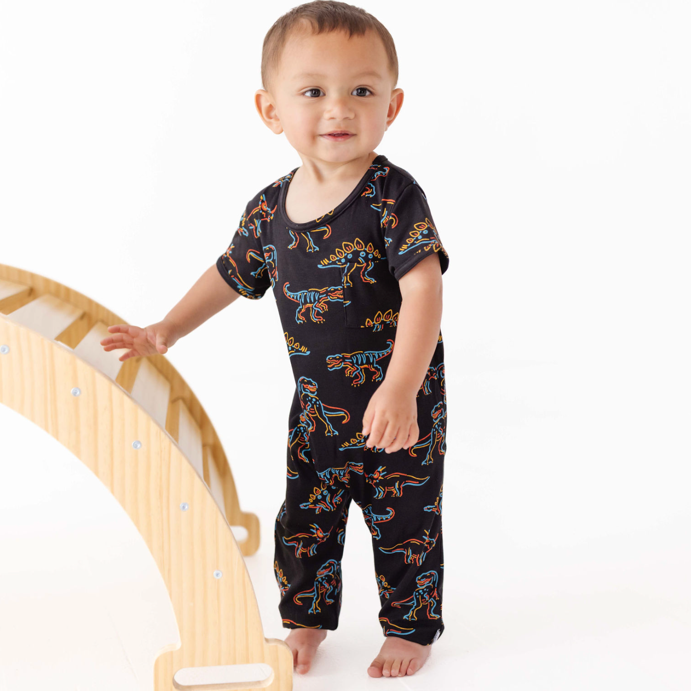 Child wearing a black dinosaur-patterned onesie standing next to a wooden climbing toy on a white background
