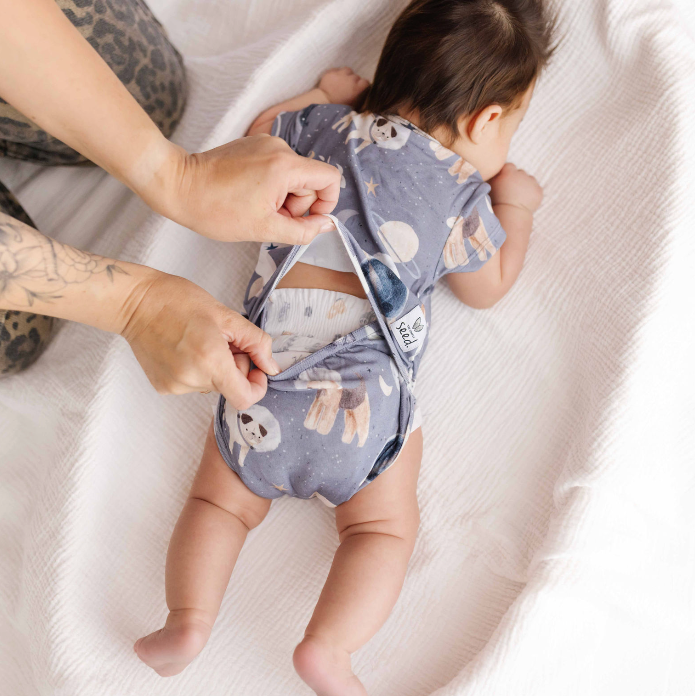 Baby wearing a patterned onesie with a person adjusting it on a white blanket.
