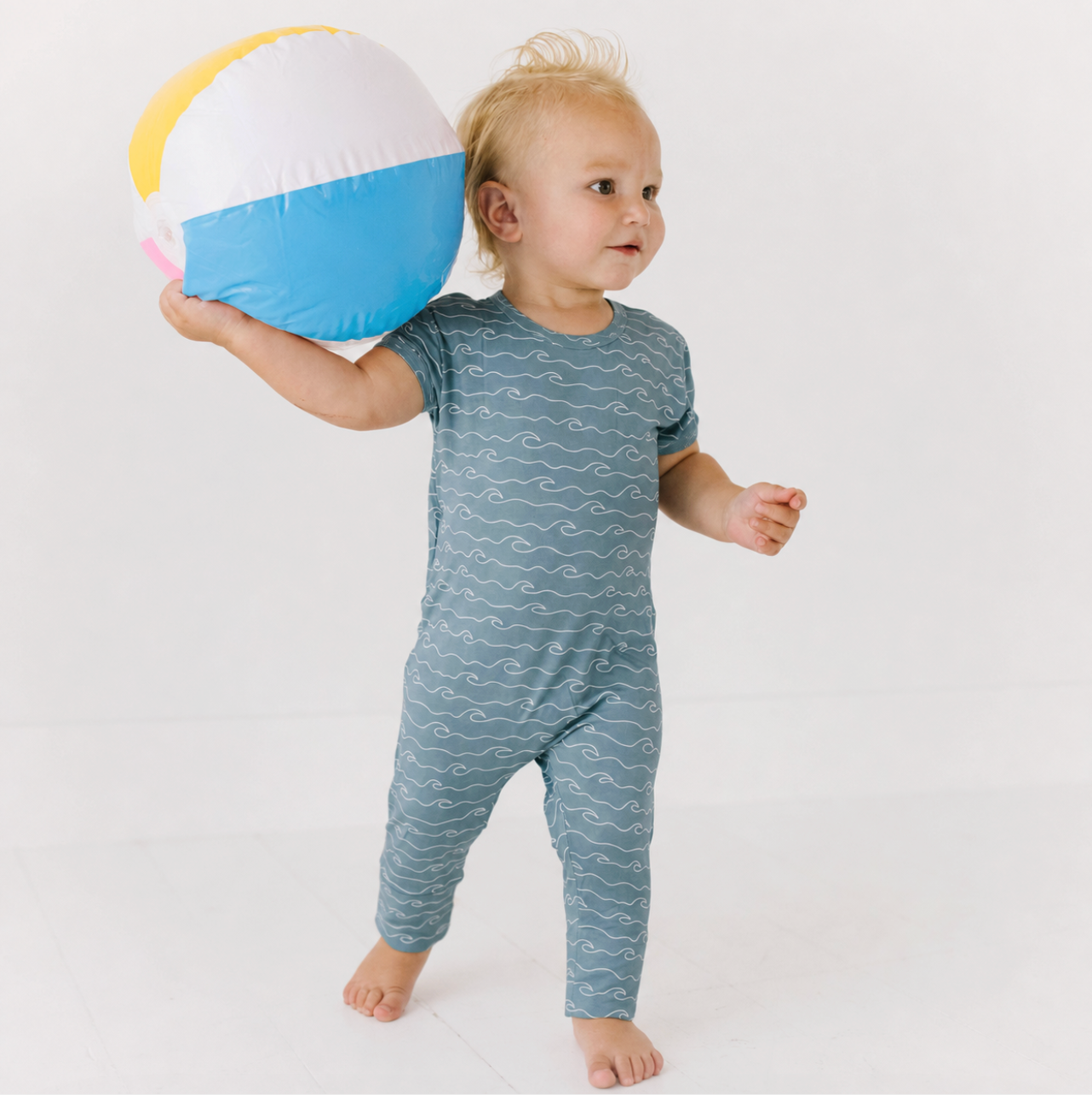 A toddler standing barefoot and holding a beach ball, wearing a short-sleeve one-piece jumpsuit in muted blue-gray with a white wavy line pattern. The background is a clean white studio setting.