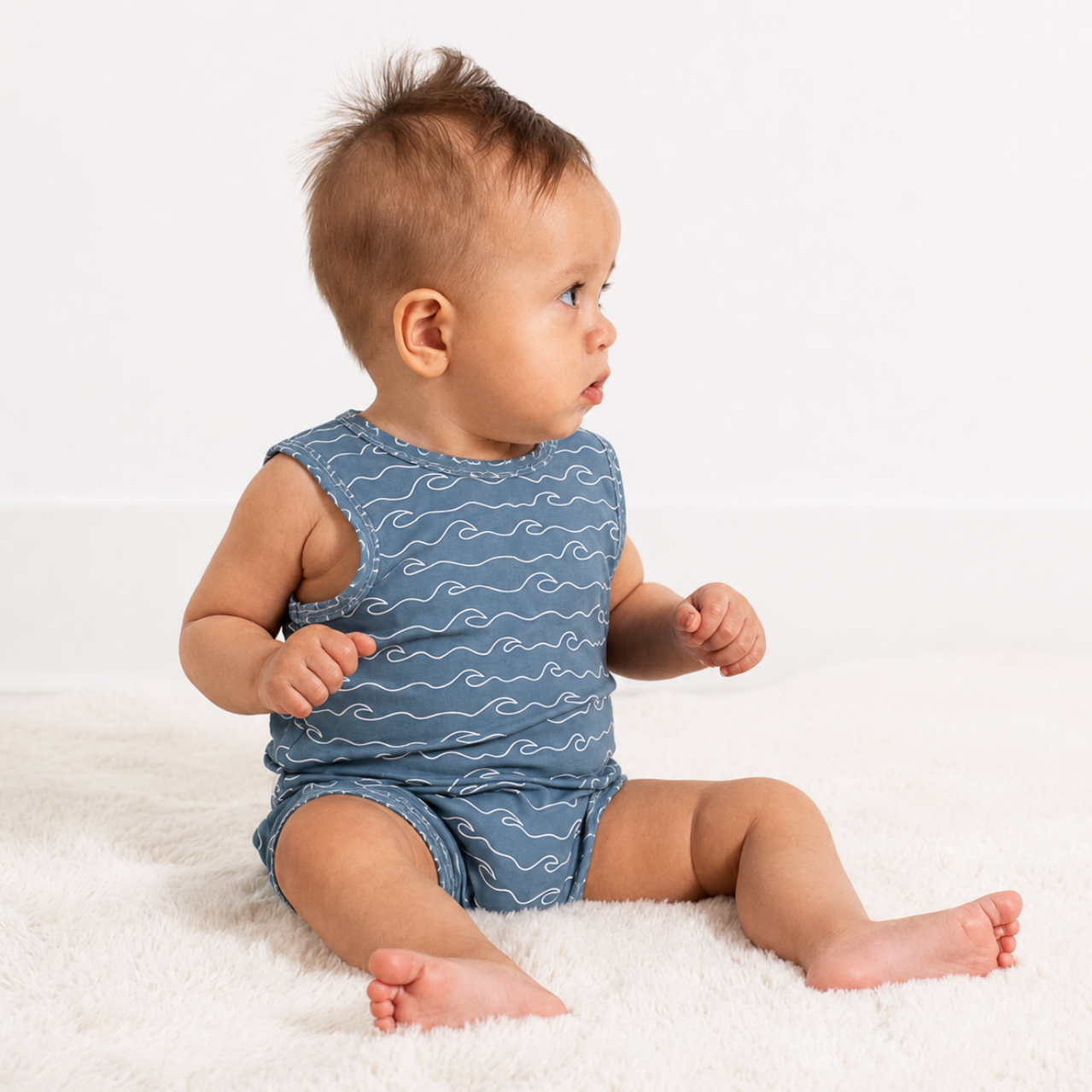 A baby sitting on a white fluffy rug wearing a sleeveless blue romper with a white wave pattern, photographed against a clean white background.