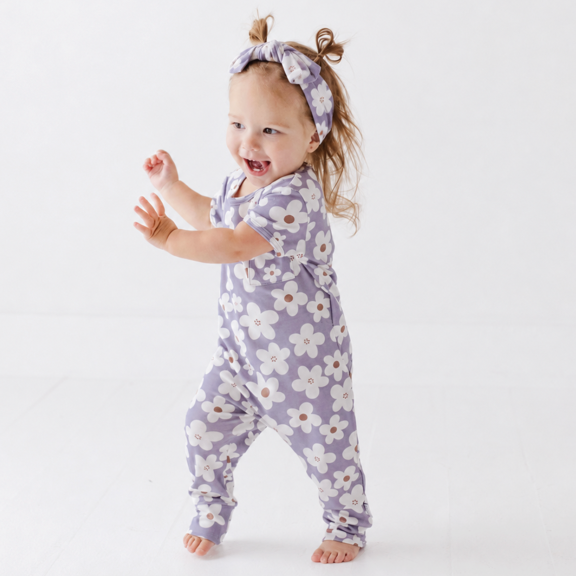A toddler girl wearing a lavender short-sleeve one-piece jumpsuit with a white and yellow floral pattern, matching headband, and small brown bow in her hair, smiling and walking on a white studio background.
