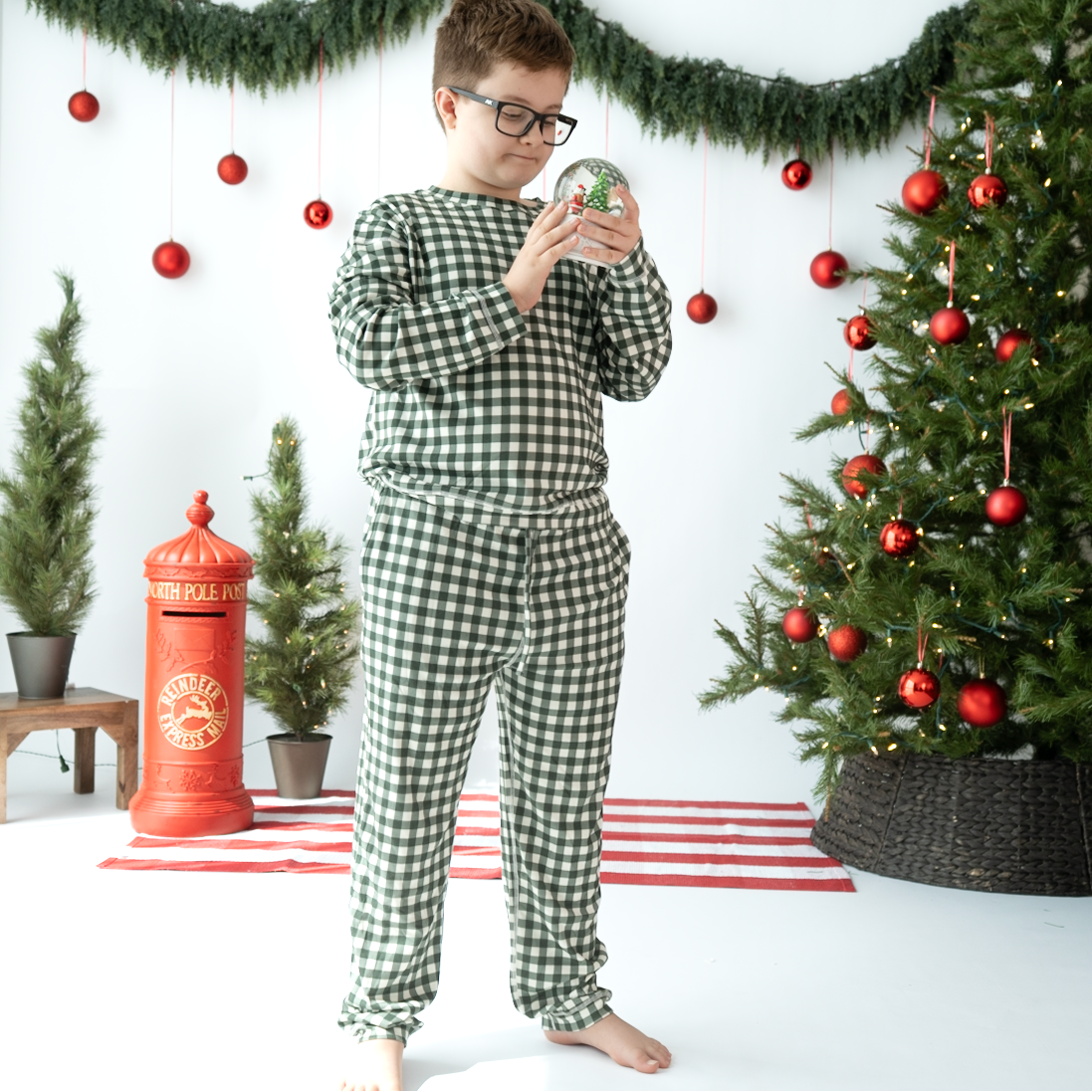 A child wearing green and white gingham check pajamas stands barefoot in a decorated Christmas room, holding a small snow globe. The scene includes a Christmas tree, garland, red ornaments, and a red post box.
