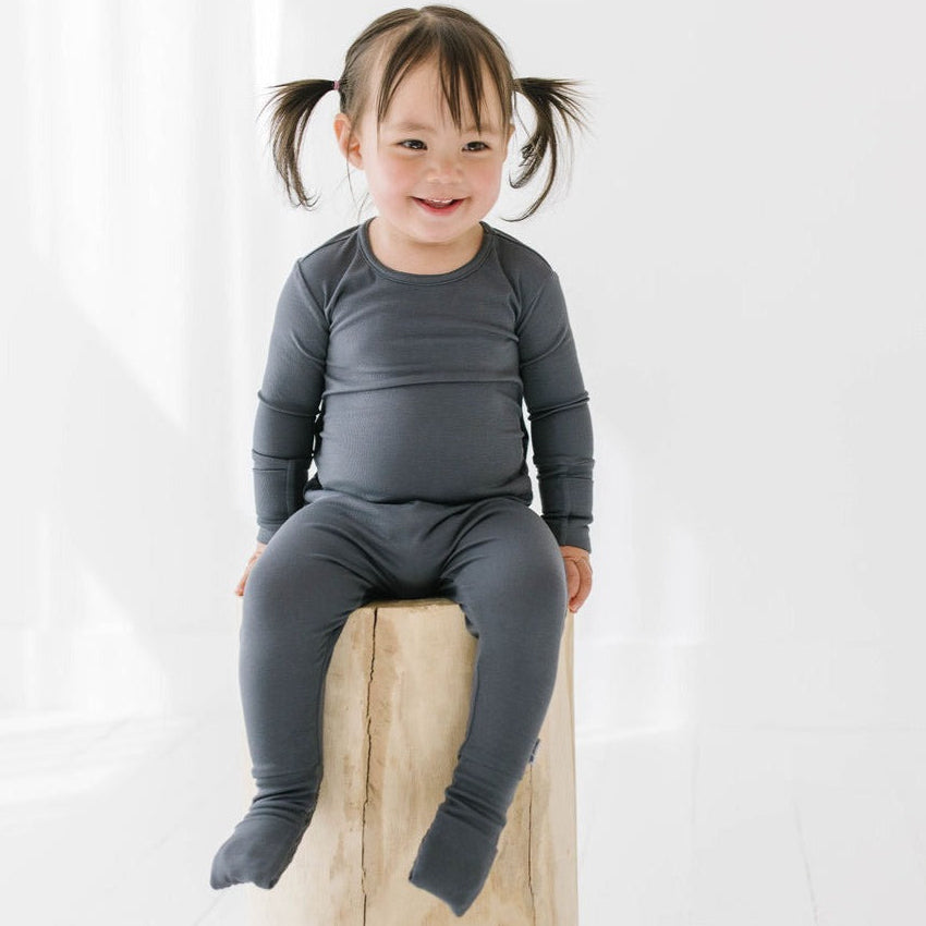A toddler sitting on a wooden stump wearing a gray bamboo romper suit with fold over mitties and footies.