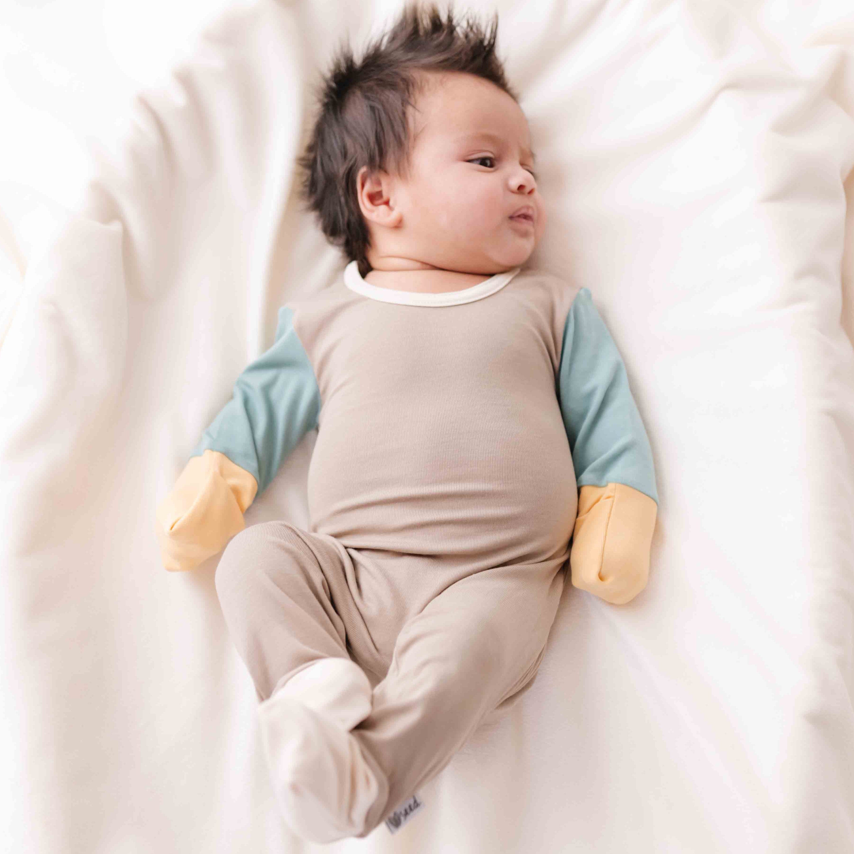 Baby lying on a white blanket wearing a beige and blue outfit.