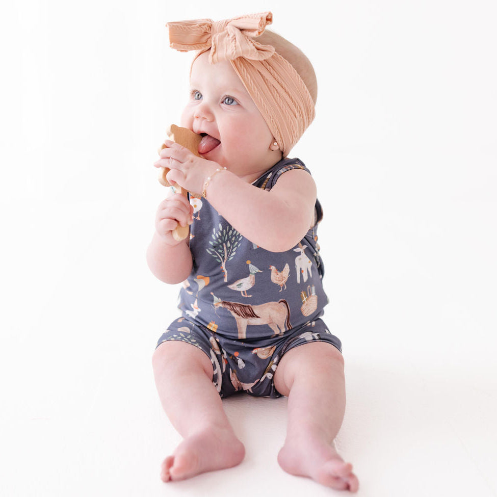Baby sitting on a white surface wearing a patterned romper and headband, holding a teething toy.