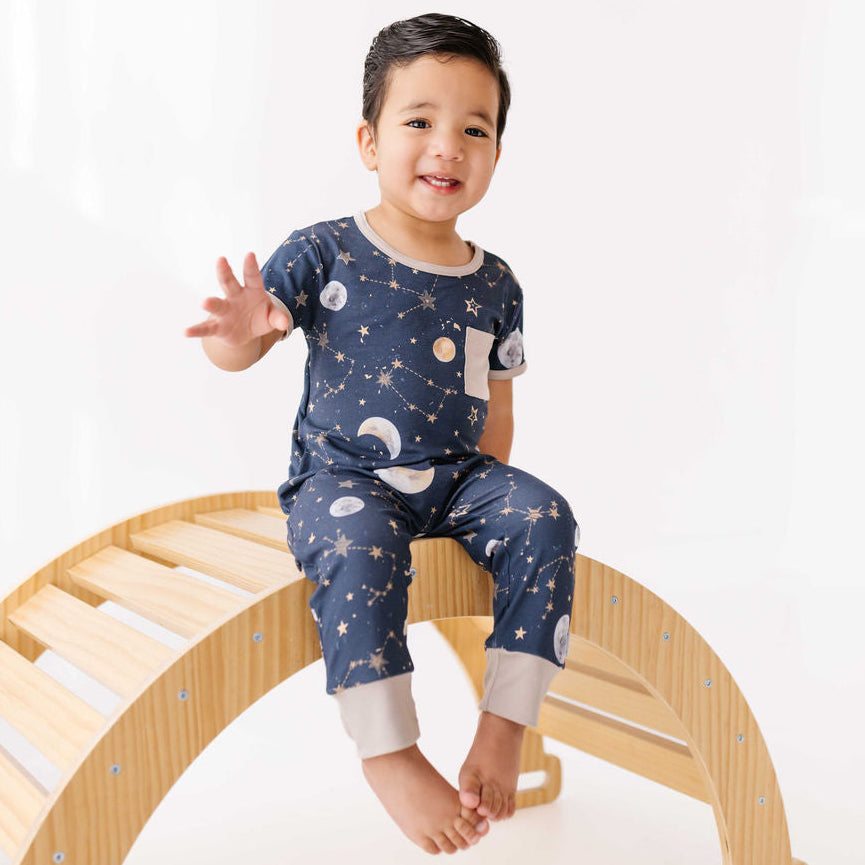 Child wearing a blue pajama set with space-themed design, sitting on a wooden arch.