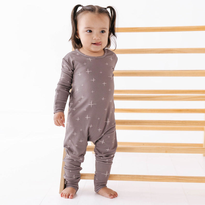 Child wearing a gray onesie with star patterns standing next to a wooden toy.