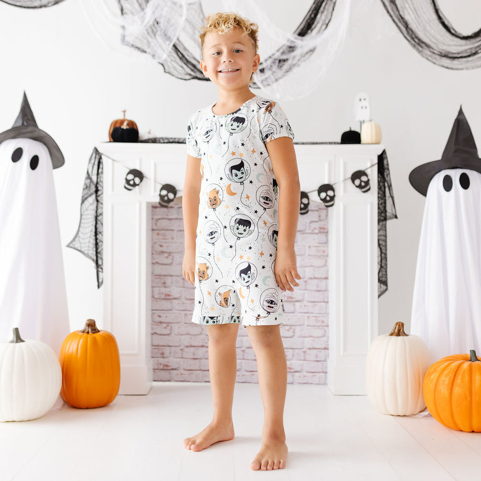 A young boy stands barefoot in front of a white fireplace mantel decorated for Halloween, wearing a short-sleeve pajama dress with a white background and a black, orange, and gray spider-and-moon print. Ghost decorations, black webbing, and pumpkins are arranged around him.