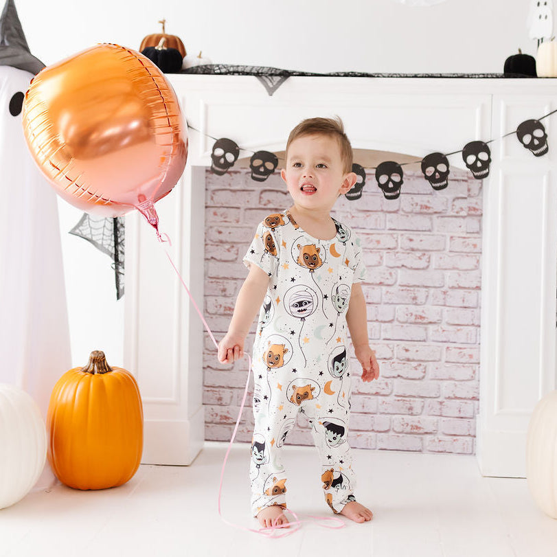 A toddler stands barefoot in a Halloween-themed room, wearing a white one-piece pajama with a cartoon animal print. The child is holding a large metallic rose-gold foil balloon, with pumpkins, black-and-white ghost figures, and a brick-patterned fireplace backdrop behind them.