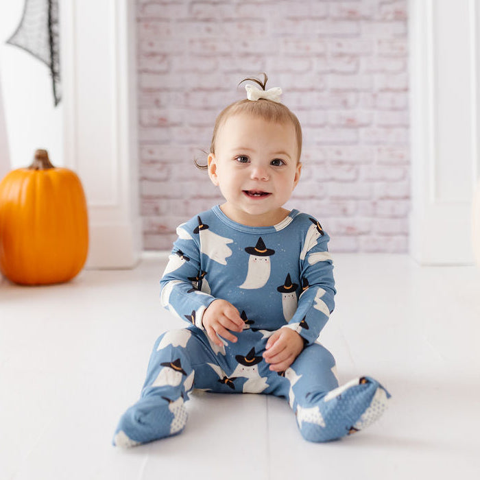 A baby sitting in front of a white fireplace mantel wearing a blue footed pajama set with a white ghost pattern. The scene is decorated for Halloween with pumpkins, ghost cutouts, and skull garland.