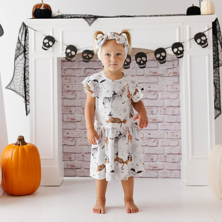 A toddler stands barefoot in front of a white fireplace mantel decorated for Halloween, wearing a white short-sleeve dress with a brown and black animal print and a matching headband. Ghost decorations, black webbing, skull garlands, and pumpkins are visible in the room.