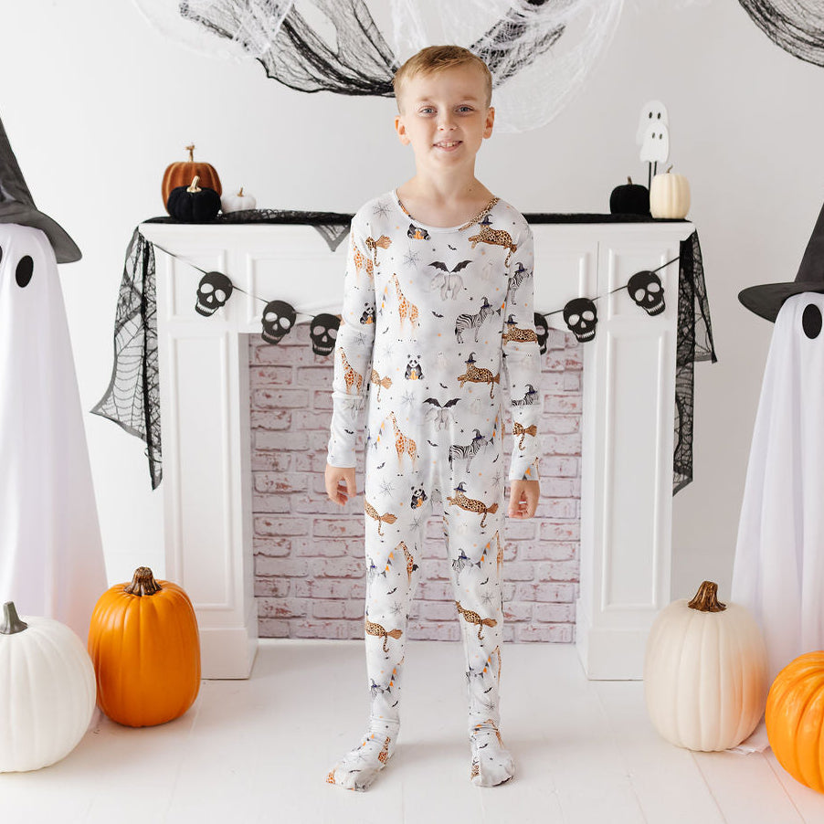A young boy stands in front of a white fireplace mantel decorated for Halloween, wearing a long-sleeve full-body pajama set with a whimsical print of cats, bats, ghosts, and pumpkins. Ghost props and pumpkins flank him on the floor and mantel.