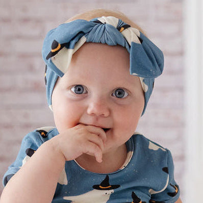 A baby sitting on a white floor wearing a blue short-sleeve romper and matching headband with a white ghost pattern, in front of a fireplace with pumpkins in the background.