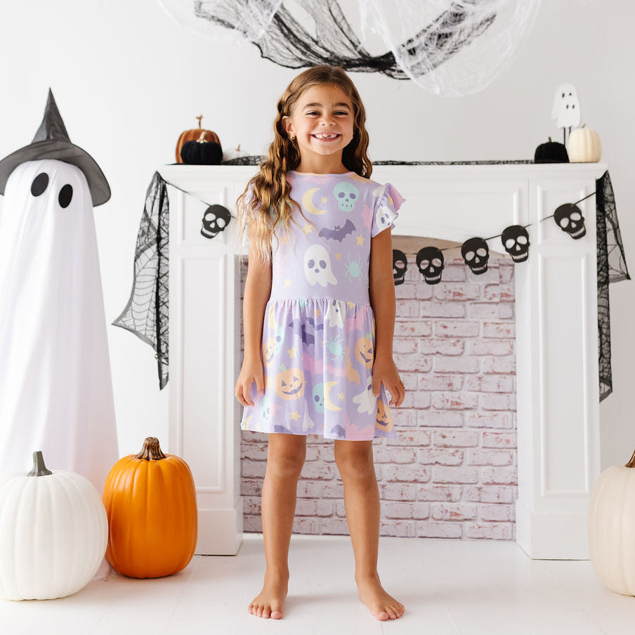 A young girl stands barefoot in front of a white fireplace mantel decorated for Halloween, wearing a short flutter-sleeve dress with a pastel multicolor print of ghosts, pumpkins, and bats. Large paper ghost props, black webbing, and pumpkins are arranged around her in a bright, festive room.