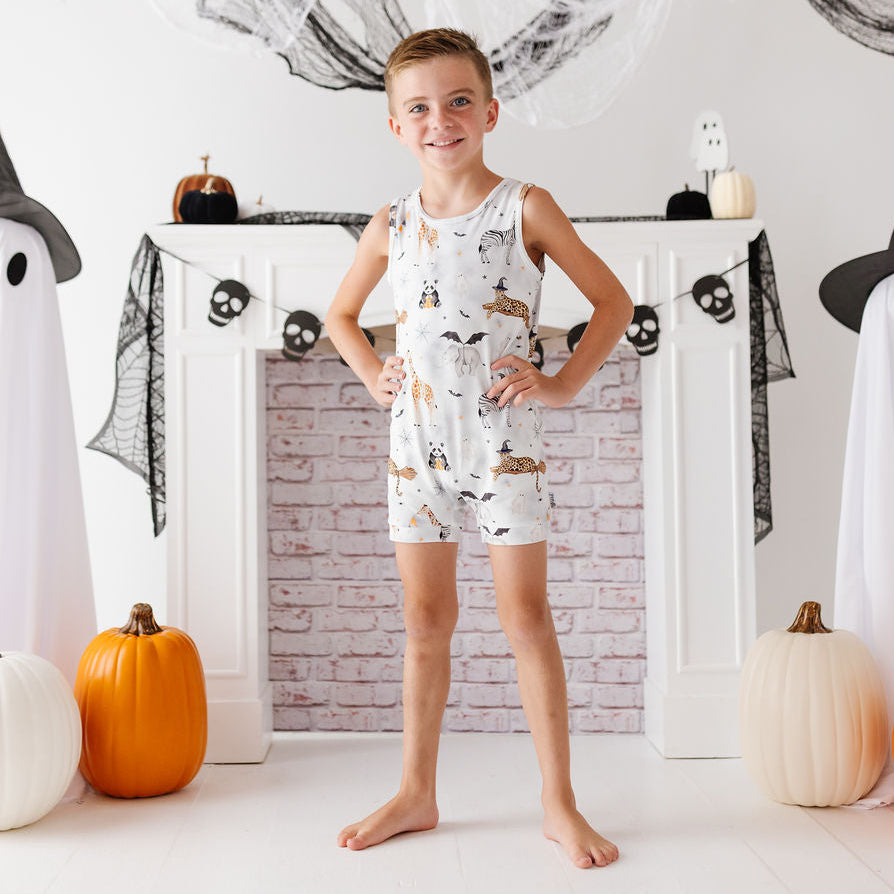 A young boy stands barefoot in front of a white fireplace mantel decorated for Halloween, wearing a white sleeveless romper with a brown and tan spider-and-bat print. Large ghost decorations, black webbing, pumpkins, and skull garlands surround him.