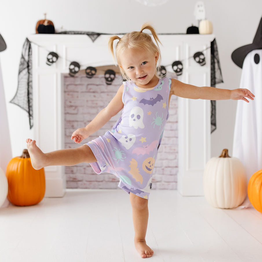 A young toddler girl with blonde pigtails poses in a Halloween-themed room, wearing a lavender sleeveless pajama dress decorated with cute ghost, pumpkin, and bat illustrations. She is balancing on one leg in front of a white brick fireplace decorated with black lace, ghost figures, and pumpkins.
